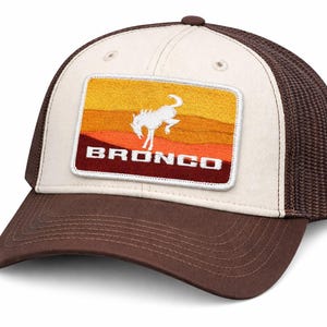 Peut inclure: Casquette de camionneur marron et beige avec un dos en filet. L'avant présente un écusson rectangulaire avec une silhouette blanche de cheval cabré sur un dégradé de coucher de soleil jaune, orange et marron. Le mot "BRONCO" est affiché en lettres blanches.