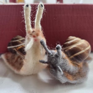 Pode incluir: Dois caracóis de feltro feitos à mão. Um caracol é principalmente branco com detalhes em castanho e bege, e o outro é cinzento com detalhes em laranja e castanho. Ambos têm conchas listradas castanhas e bege e antenas longas e direitas.