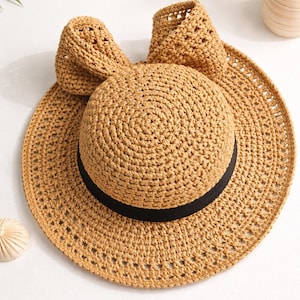 May include: A woven straw hat with a wide brim and a black band. The hat features a rounded crown and a decorative bow on the back. The hat is a light brown color and is suitable for sun protection.