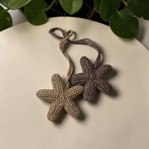 May include: Two crocheted star-shaped ornaments in beige and grey-purple. Each star has five points and is connected to a twisted cord for hanging. The ornaments are handmade and have a textured appearance.