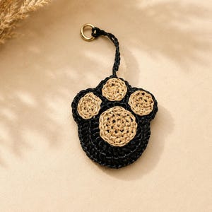 May include: A crocheted animal paw keychain featuring a black base with three small tan circular pads and one large central tan pad. The keychain includes a black crocheted loop attached to a small gold-toned metal ring for easy attachment.