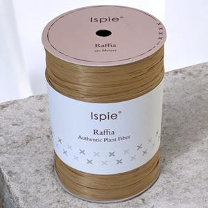 May include: A spool of Kraft-colored raffia ribbon. The label reads "Ispie Raffia Authentic Plant Fiber" and "250 Meters". The word "Kraft" is printed at the bottom. The spool is sitting on a gray surface.