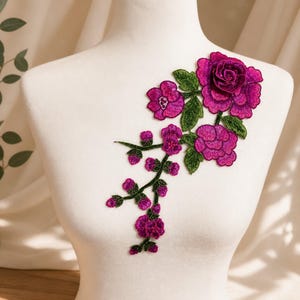 Puede incluir: Un parche bordado floral que presenta rosas de color magenta vibrante y morado intenso con hojas y tallos verdes detallados. El diseño consta de varias flores en plena floración y pequeños capullos dispuestos en un patrón de rama en cascada.