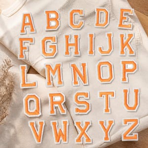Puede incluir: Una colección de veintiséis parches termoadhesivos individuales que muestran las letras de la A a la Z en una fuente de estilo universitario. Cada letra está bordada con hilo naranja brillante y delineada en blanco, creando un aspecto limpio y contrastado.