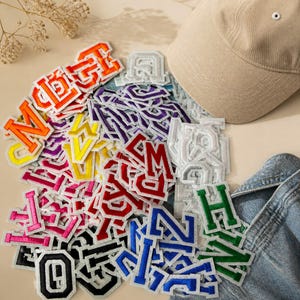 Könnte beinhalten: Eine Sammlung von Buchstaben-Aufnähern im College-Stil in verschiedenen Farben wie Orange, Gelb, Pink, Rot, Lila, Blau, Grün, Schwarz und Weiß, angeordnet neben einer beigen Baseballkappe und einem Stück blauem Jeansstoff.