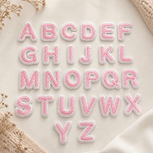 Puede incluir: Un conjunto completo de veintiséis parches bordados del alfabeto, cada uno con una letra rosa y un borde blanco. Las letras de la A a la Z están dispuestas en cuatro filas, mostrando una textura cosida uniforme y un estilo de fuente redondeado.