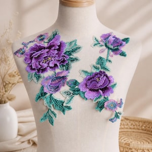 May include: A floral embroidery patch featuring large, vibrant purple peony blossoms with intricate dark purple shading and detailed green leaves. The design consists of two connected floral clusters with stems and smaller buds, showcasing dense thread work and rich color gradients throughout the entire embroidered textile piece.