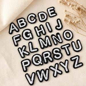 Puede incluir: Un conjunto de veintiséis parches termoadhesivos individuales que muestran las letras del alfabeto de la A a la Z. Cada letra está representada en una fuente sans-serif blanca y negrita con un borde bordado negro grueso y definido.