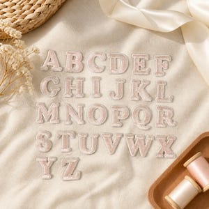May include: A complete set of twenty-six alphabet letters as individual iron-on patches. Each letter features a soft, light pink velvet-like texture with a stitched border. The letters are arranged in rows on a neutral fabric surface.