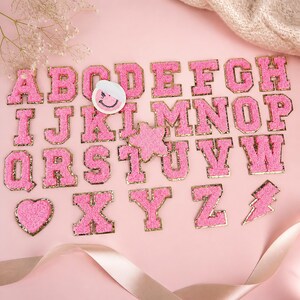 Può includere: Una collezione di toppe a lettera in ciniglia rosa con bordi glitterati dorati, disposte dalla A alla Z. Il set include una toppa circolare con faccina sorridente, una stella rosa, un cuore e un fulmine, tutti in tessuto ciniglia.