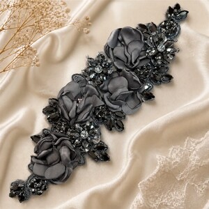 May include: A decorative appliqué featuring dark grey fabric flowers and black faceted crystals arranged in an elongated floral pattern. The piece includes layered satin-like petals and clusters of dark, reflective beads on a dark mesh backing.