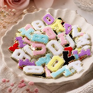 Puede incluir: Una colección de pequeños parches de letras bordadas y difusas en varios colores pastel y llamativos, incluyendo blanco, rosa, azul claro, amarillo, morado y negro. Cada letra presenta una superficie texturizada tipo felpa y un borde dorado.