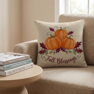 Könnte beinhalten: Ein quadratisches Zierkissen mit einem strukturierten beigen Stoffbezug, bedruckt mit einer Illustration von drei orangefarbenen Kürbissen, umgeben von dunkelroten und grünen Herbstblättern. Der Text Fall Blessings steht in dunkelbrauner Schreibschrift.
