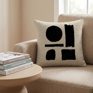 Puede incluir: Un cojín cuadrado con una funda de tela beige texturizada que presenta un estampado gráfico negro. El diseño consta de un círculo sólido, un rectángulo vertical, una barra horizontal, una forma redondeada y un cuadrado, dispuestos en una composición equilibrada.