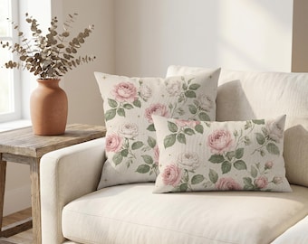Puede incluir: Dos cojines decorativos con un estampado botánico de rosas rosas y blancas con hojas verdes sobre un fondo beige claro. Los cojines cuadrados y rectangulares están decorados con pequeños detalles de estrellas doradas sobre el diseño floral.