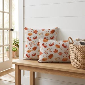 May include: Two square throw pillows featuring a repeating pattern of orange and yellow autumn leaves and clusters of red berries on a light cream background. The pillows are placed on a wooden surface next to a woven wicker basket.