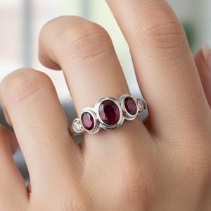 Könnte beinhalten: Ein silberner Ring mit drei ovalen, tiefroten Edelsteinen und kleineren, runden, klaren Steinen. Der Ring befindet sich an einem Finger und unterstreicht sein elegantes Design. Der Ring ist ein Schmuckstück. Die Edelsteine sind in einem Silberband gefasst.