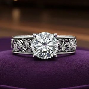 Könnte beinhalten: Ein silberfarbener Ring mit einem großen, runden, klaren Mittelstein im Brillantschliff. Die Ringschiene ist mit komplizierten, erhabenen Schnörkelmustern verziert, die sich um den Umfang erstrecken, ergänzt durch ein feines Perlenranddetail.
