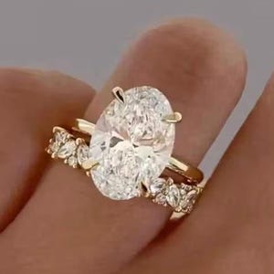 Könnte beinhalten: Ein Goldring mit einem großen, oval geschliffenen Mittelstein, der von vier Krappen gehalten wird, flankiert von einer Ringschiene, die mit mehreren kleineren, marquise-geschliffenen Steinen besetzt ist. Die Ringschiene ist poliert.