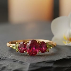 Könnte beinhalten: Ein goldener Ring mit drei ovalen, roten Edelsteinen und zwei kleineren grünen Edelsteinen. Der Ring liegt auf einer dunkelgrauen Oberfläche, mit einer weißen Blume und zwei brennenden Kerzen im Hintergrund.