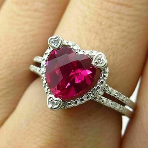 May include: A silver-toned ring featuring a large, faceted, heart-shaped deep pink gemstone as the centrepiece. The ring is surrounded by a halo of small, clear, sparkling stones, with additional small stones set along the split metal band.