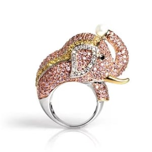 Könnte beinhalten: Ein silberfarbener Ring mit einem elefantenförmigen Schmuckelement, das mit rosa und gelben Kristallen besetzt ist. Der Elefant hat eine weiße Perle an der Rüsselspitze, ein kleines schwarzes Auge und einen goldfarbenen Stoßzahn auf einer Ringschiene.