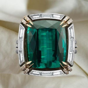 Könnte beinhalten: Ein Ring mit einem großen, tiefgrünen Edelstein im Kissenschliff in der Mitte. Der Stein ist von einem Rahmen aus rechteckigen Baguette-Diamanten umgeben und an den vier Ecken mit kleinen, in Gold gefassten runden Diamanten verziert.