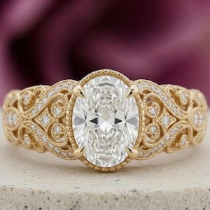 Könnte beinhalten: Ein goldener Verlobungsring mit einem oval geschliffenen Diamanten. Das Band weist aufwendige Filigranarbeiten und kleine Akzentdiamanten auf. Der Ring liegt auf einer neutralen Oberfläche mit verschwommenem lila Hintergrund.