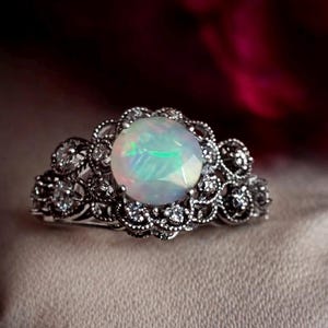 May include: A silver ring featuring a large, round opal gemstone with iridescent colors. The opal is surrounded by a detailed, ornate setting with smaller, clear gemstones. The ring has a vintage or antique style.