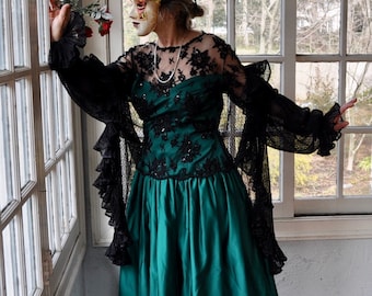 May include: A woman wearing a green satin gown with a black lace overlay and a masquerade mask. The gown has a fitted bodice and a full skirt. The woman is standing in front of a window.
