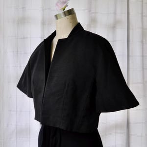 May include: A black cropped jacket with short, wide sleeves and a textured, ribbed fabric finish. The jacket features an open front design with a structured collarless neckline, providing a clean and simple silhouette that sits at the waist.