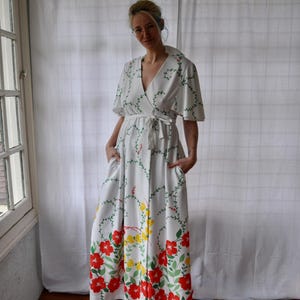 May include: White wrap dress with a floral pattern. The dress features a V-neck, short sleeves, and a tie waist. Red and yellow flowers with green leaves adorn the bottom of the dress. The dress has pockets.