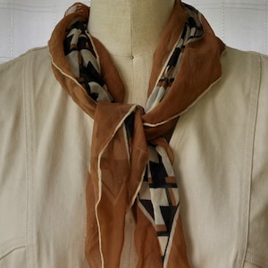 May include: A brown scarf with a geometric pattern in black, white, and tan is draped around a mannequin's neck. The scarf is knotted at the front and has a delicate, sheer appearance. A light pink rose sits atop the mannequin's head.