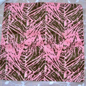 May include: A square, pink fabric with a brown, abstract pattern. The design features irregular, dark brown shapes against a light pink background, with thin white vertical lines. The fabric is likely a scarf or a similar textile.