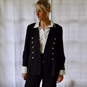 May include: Black pinstripe blazer with gold buttons, paired with matching pinstripe pants. The blazer features a double-breasted design and is worn over a white collared shirt. The outfit is a classic, tailored look.