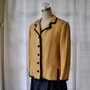 May include: A mustard yellow blazer with black trim and buttons. The blazer is displayed on a mannequin. The jacket has a classic design with a notched collar and long sleeves. The blazer is made of a textured fabric.