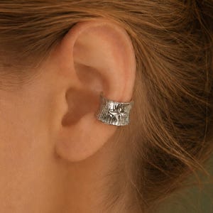 May include: A silver ear cuff featuring a textured, bark-like surface and a small, embossed dragonfly motif centred on the band. The metallic accessory wraps around the cartilage of the ear, showcasing a polished finish with intricate detailing.