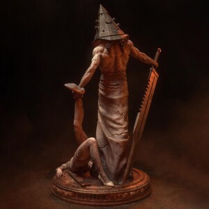 May include: A detailed statue of a figure with a pyramid-shaped head, holding a large blade and a smaller figure. The statue is primarily brown and tan, with a textured appearance. The base is circular and detailed.