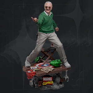 Puede incluir: Estatua de Stan Lee con suéter verde y pantalón gris, sobre una base con logotipos de Marvel Comics y elementos de cómic. La base incluye una máscara de Spider-Man, puños de Hulk y un escudo del Capitán América.