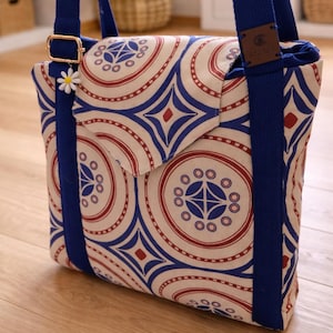 May include: A patterned crossbody bag with a flap closure. The bag features a repeating design of red, blue, and cream-colored circles and geometric shapes. It has a blue strap and a small daisy charm.