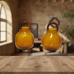 Puede incluir: Dos jarrones de cristal color ámbar con cuerpo redondeado y asa marrón oscuro. Los jarrones tienen el interior amarillo y se exhiben sobre una superficie de madera. El fondo es un interior borroso con una ventana.