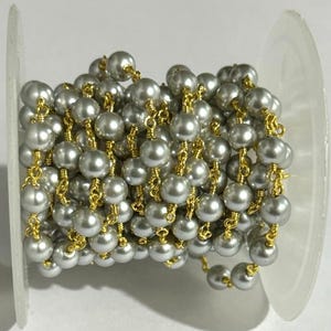 Puede incluir: Una longitud continua de cuentas de perlas gris plateado unidas por un fino alambre de metal dorado, enrollada alrededor de un carrete de plástico transparente. Las cuentas esféricas son uniformes en tamaño y color, creando eslabones dorados.