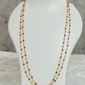 Peut inclure: Un collier doré composé de deux rangées de petites perles rouges et blanches. Le collier est présenté sur un mannequin blanc. Les perles sont espacées régulièrement le long de la chaîne dorée, créant un design délicat. Le fond est de couleur neutre.