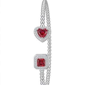 Può includere: Un braccialetto d'argento con un design a perline. Presenta una gemma rossa a forma di cuore e una gemma rossa quadrata, entrambe circondate da piccole pietre trasparenti. Il braccialetto è un gioiello.