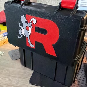 Pode incluir: Uma caixa de armazenamento retangular preta com dois fechos de bloqueio vermelho brilhante no topo. A frente da caixa apresenta uma letra R vermelha grande e um gráfico cinzento e branco da personagem Mewtwo na superfície preta.