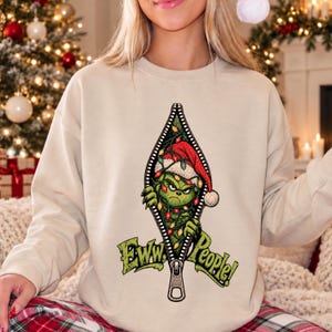May include: A beige crewneck sweatshirt featuring a graphic of a green creature wearing a red Santa hat, tangled in colorful Christmas lights, peeking through a vertical zipper opening with the text Eww, People! printed in green below.