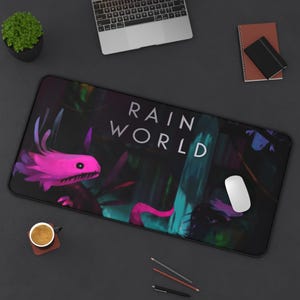 May include: A black rectangular desk mat featuring the words "RAIN WORLD" in white. The mat has a colorful illustration of a pink creature and a forest scene. A white computer mouse sits on the mat.