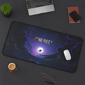 Puede incluir: Alfombrilla de ratón rectangular negra con el texto "ONE PIECE" en blanco y rojo. El diseño incluye un cielo nocturno púrpura y azul con una luna llena y una silueta de personaje. Un ratón blanco y otros útiles de oficina están sobre la superficie oscura.