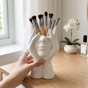 Könnte beinhalten: Ein weißer Keramik-Make-up-Pinselhalter in Form eines Gesichts mit Händen, die die Augen bedecken. Der Halter ist mit verschiedenen Make-up-Pinseln gefüllt. Der Halter steht auf einer Holzoberfläche.