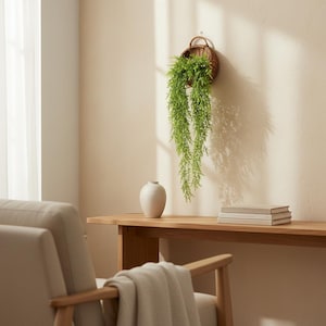 Könnte beinhalten: Ein geflochtener Korb mit einer herabhängenden grünen Pflanze hängt an einer beigefarbenen Wand. Eine kleine weiße Vase und zwei Bücher stehen auf einem Holztisch. Ein beigefarbener Sessel befindet sich im Vordergrund.